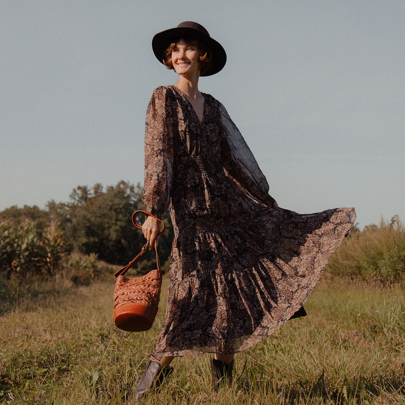 A smiling woman in a field carrying a woven leather crossbody bag