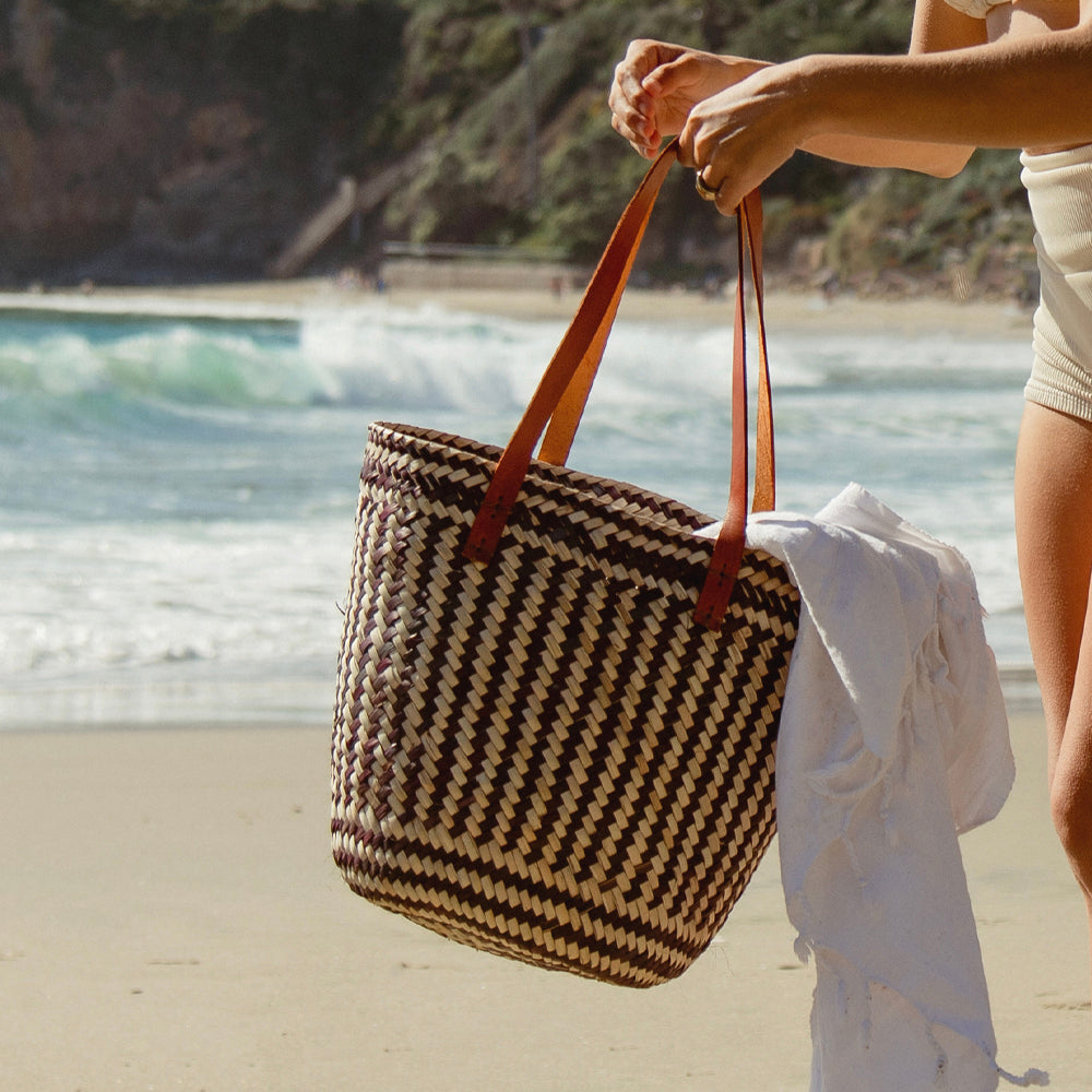 Woman holding black and white woven straw tote bag by ocean waves