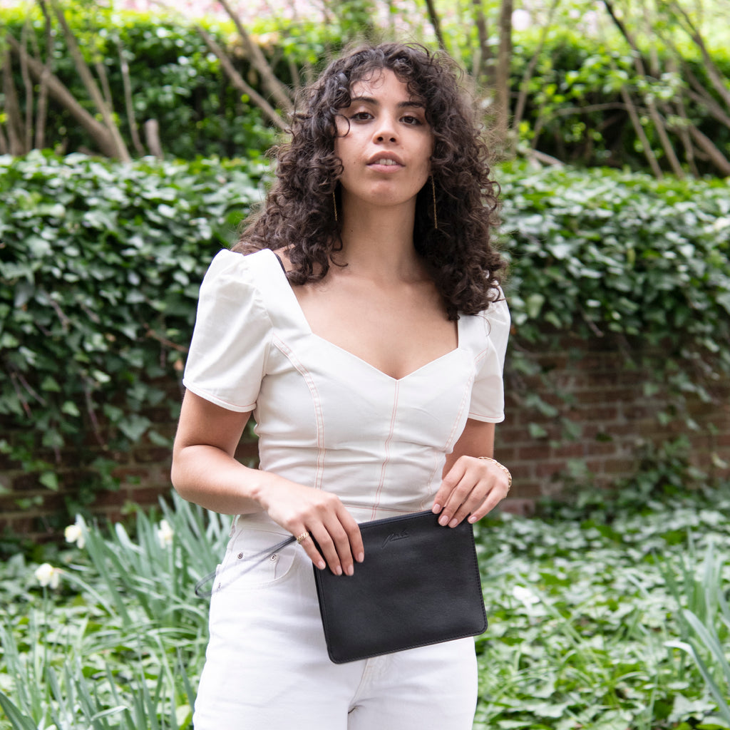 Young woman in white top and pants holding a black leather wristlet pouch clutch